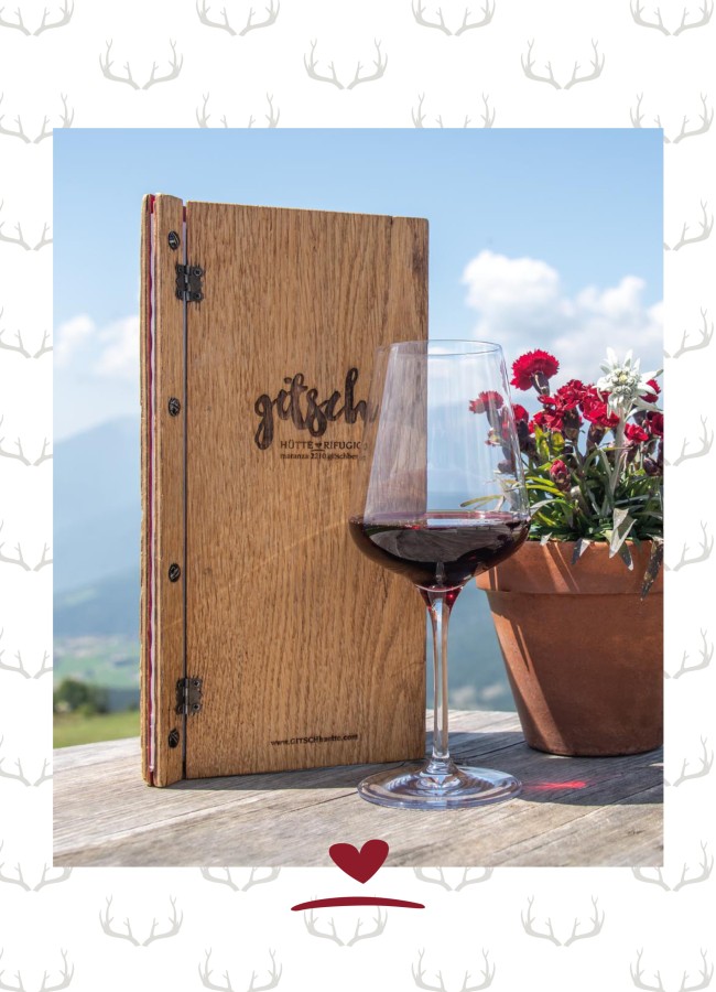 Menükarte aus Holz neben einem Topf mit Blumen und einem Glas Rotwein auf einem Holztisch. In Hintergrund Berge und blauer Himmel.