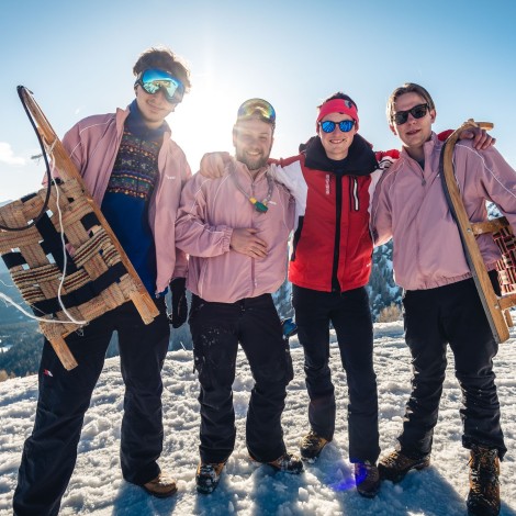 Four people in winter clothing on a snow-covered mountain. Two of them are holding sledges in their hands. In the background, there is a clear sky and snow-covered mountains.