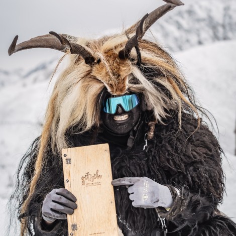 A person dressed in a Krampus costume with horns, fur and a mask in a snowy landscape. The person is holding a wooden menu card in their hand and pointing at it.