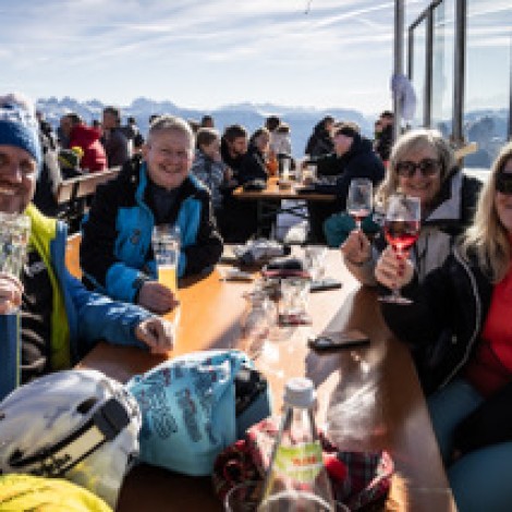 Four people are sitting at a table on a sunny terrace with drinks in their hands, and in the background you can see snow-capped mountains under a blue sky.