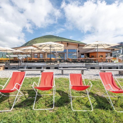 Four red deckchairs with white metal frames stand on a lawn in front of a terrace with parasols and a wooden building in the mountains.