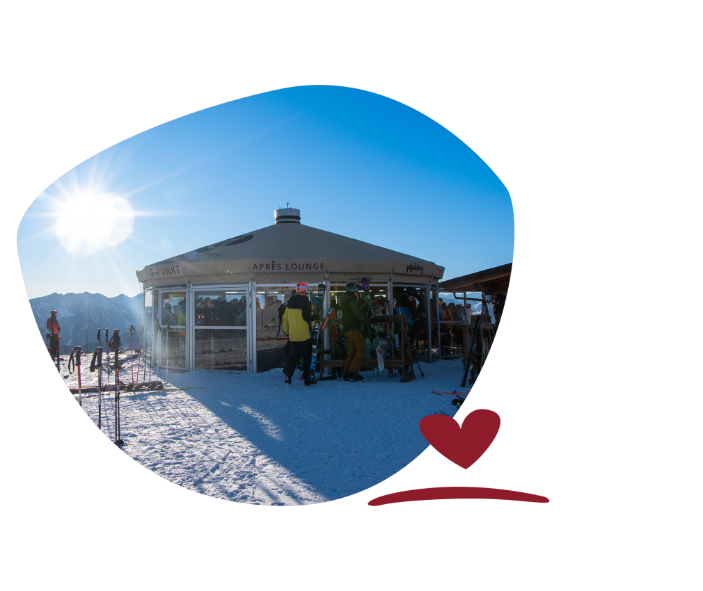 Skiers in front of the après-lounge, a round building with a glass front, on a snow-covered mountain in the sunshine.
