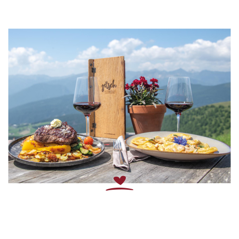 Eine Menükarte aus Holz und zwei Gerichte mit zwei Weingläsern Rotwein auf einem Holztisch im Freien. In Hintergrund Berge und blauer Himmel.