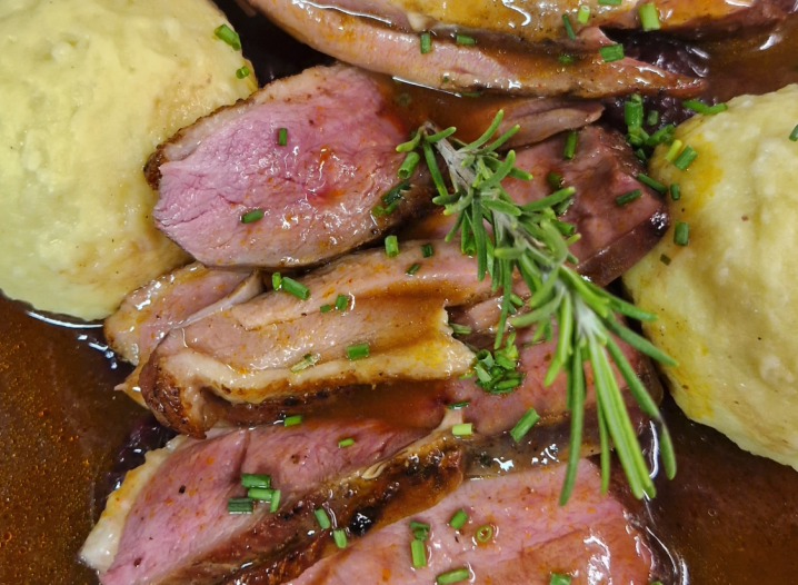 On a plate are several slices of pink roasted lamb loin with gravy, accompanied by two dumplings and a sprig of rosemary as garnish.