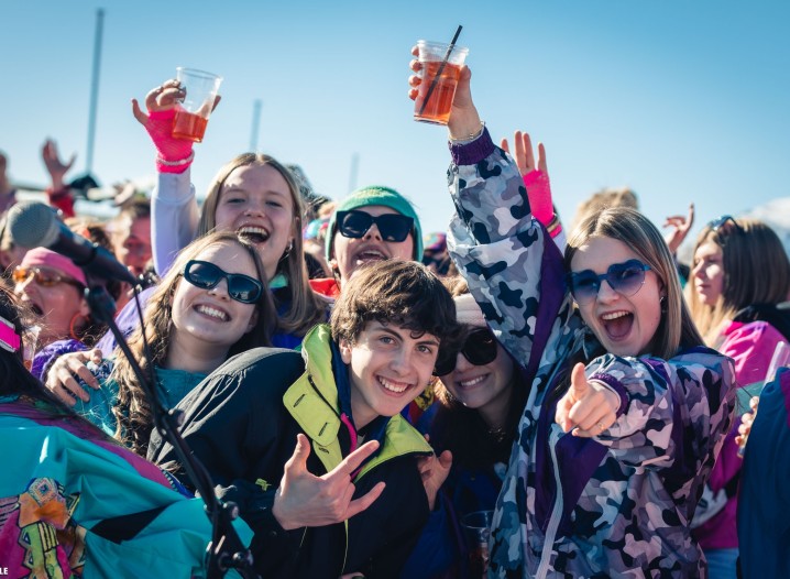 A group of young people in colourful winter clothing are celebrating outdoors, holding drinks in their hands.