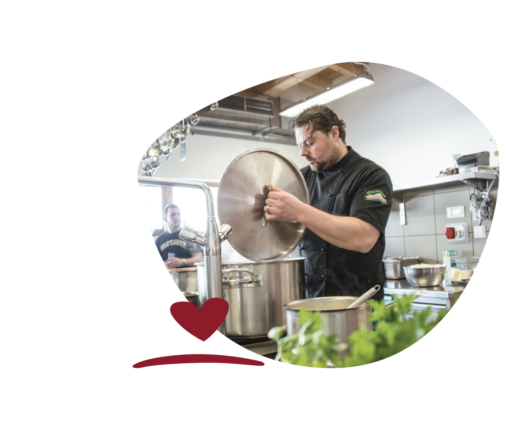 Der Chefkoch in der Küche beim Kochen. Im Vordergrund Kräuter und ein Topf.