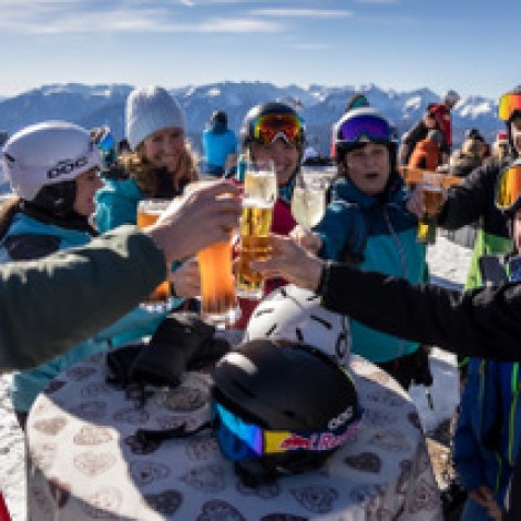Several people in ski gear toast with their drinks in front of a high table in the snow. Mountains and blue sky in the background.