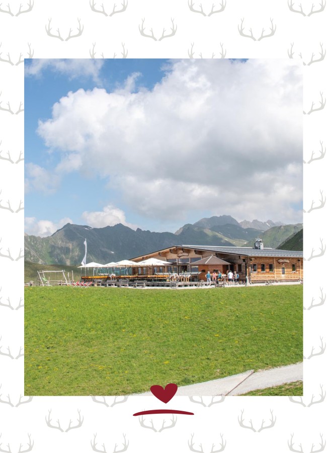 Bergrestaurant mit Terrasse umgeben von grünen Wiesen und einem Wanderweg. In Hintergrund ein Bergpanorama und ein leicht bewölkter Himmel.