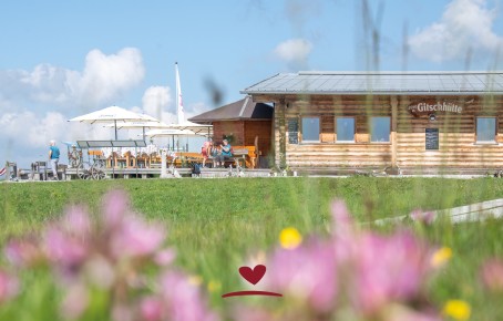 A mountain restaurant with a large terrace with many tables and parasols, surrounded by green meadows and blue skies.