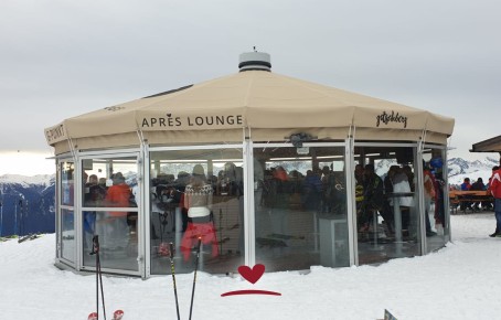 Viele Menschen in einer von Glas umrundete Après-Lounge mit einem Schirmdach auf der Skipiste mit Berge im Hintergrund.