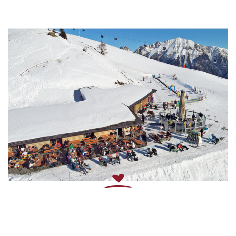 Snow-covered mountain hut with a terrace where several people are sitting, with snow-capped mountains and ski lifts with cable cars in the background.