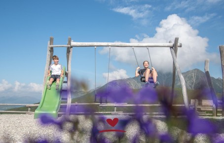 Two children on the swing and slide, with mountains and blue sky in the background.