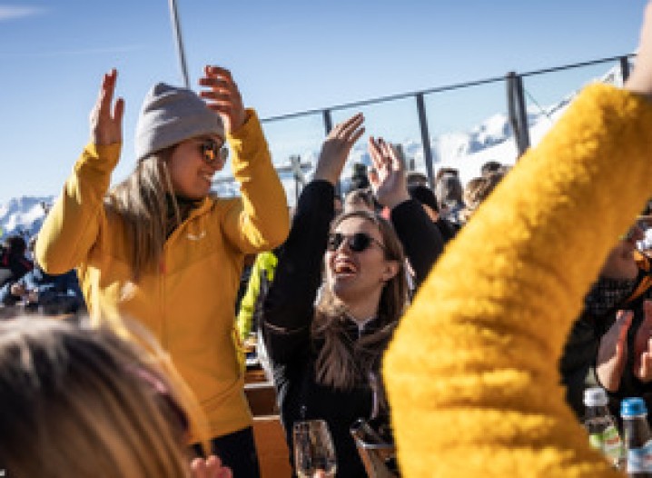 Two happy women dancing on a terrace, clapping their hands. In the background, several people sitting at tables, a snow-covered mountain landscape and a blue sky.