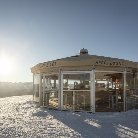 An après-lounge next to a mountain hut surrounded by snow, mountains and a blue, sunny sky.