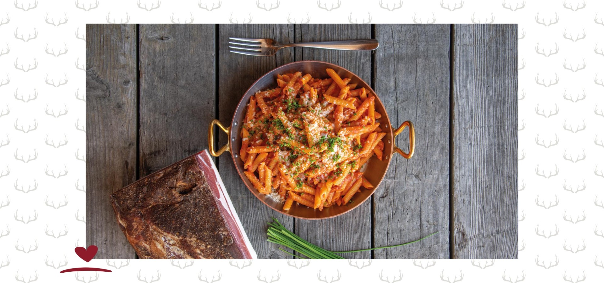 Eisenpfanne mit Nudeln und Tomatensauce auf einem Holztisch. Daneben eine Gabel, ein Stück Speck und Schnittlauch.