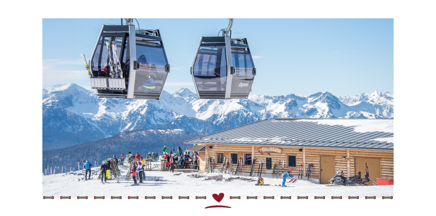 Two cable car cabins float above a snow-covered mountain station with several skiers and mountain peaks in the background.
