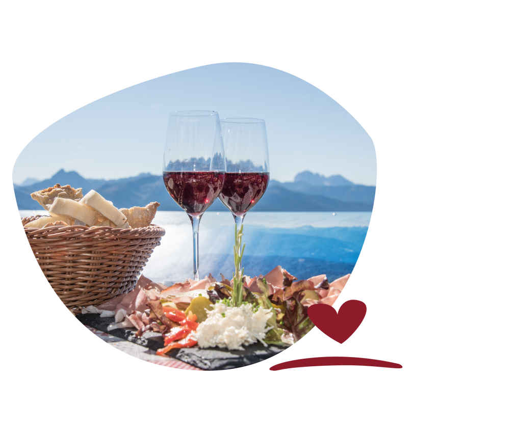 Two glasses of red wine and a basket of bread on a table with a plate of cold cuts and cheese. Mountains and blue sky in the background.