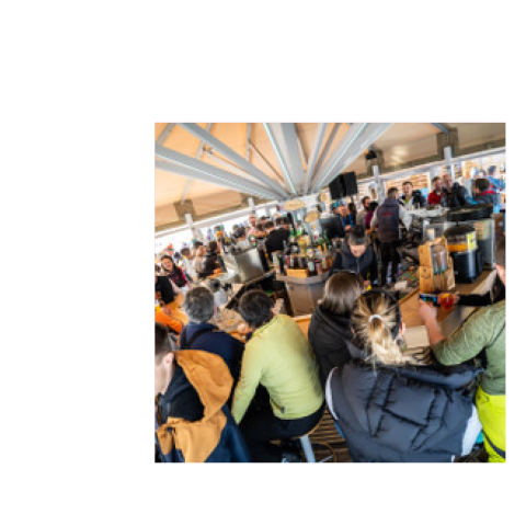 Several people are sitting and standing at a bar in a bright, covered room with exposed ceiling beams.