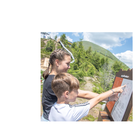Two children point to a brown information board in a green mountain landscape with blue sky.