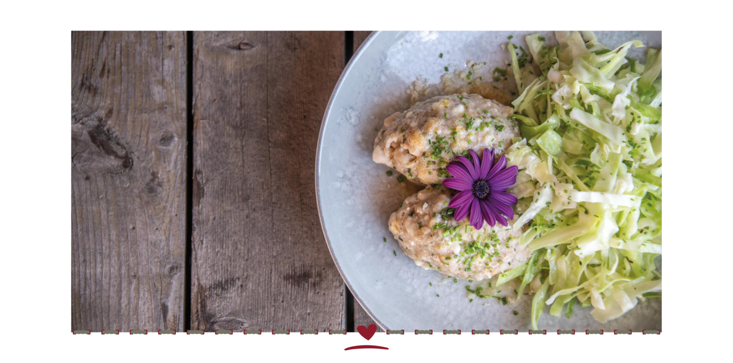 Weißer Teller mit zwei Käseknödel und Krautsalat auf einem Holztisch. Als Dekoration eine violette Blume.