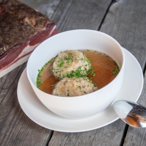 A bowl with two herb dumplings on a wooden table. In the background, a piece of speck.