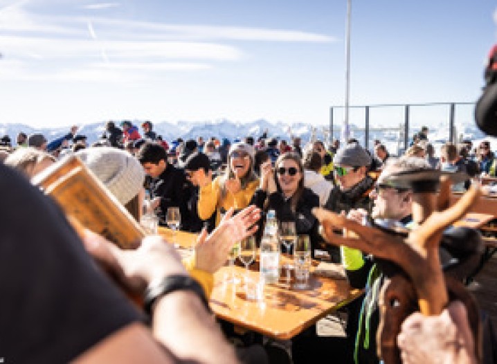 Many people in winter clothing are seated at tables on a terrace in front of a mountain restaurant. Surrounded by snow-covered mountains and blue skies.