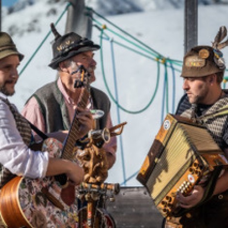 Three men playing two accordions and a guitar. In the background, snow-capped mountains and a climbing frame for children.