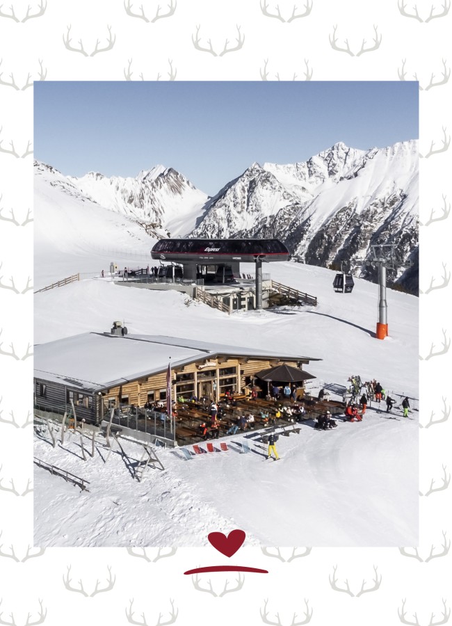 Bergrestaurant mit Terrasse und Sonnenschirmen auf schneebedecktem Berg, im Hintergrund schneebedeckte Gipfel und blauer Himmel.