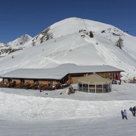 A mountain restaurant surrounded by a snowy landscape. In the background, a cable car and snow-covered mountains under a blue sky.