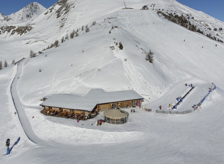 A mountain restaurant surrounded by a snowy landscape and a small ski lift for children. In the background, snow-capped mountains under a blue sky.