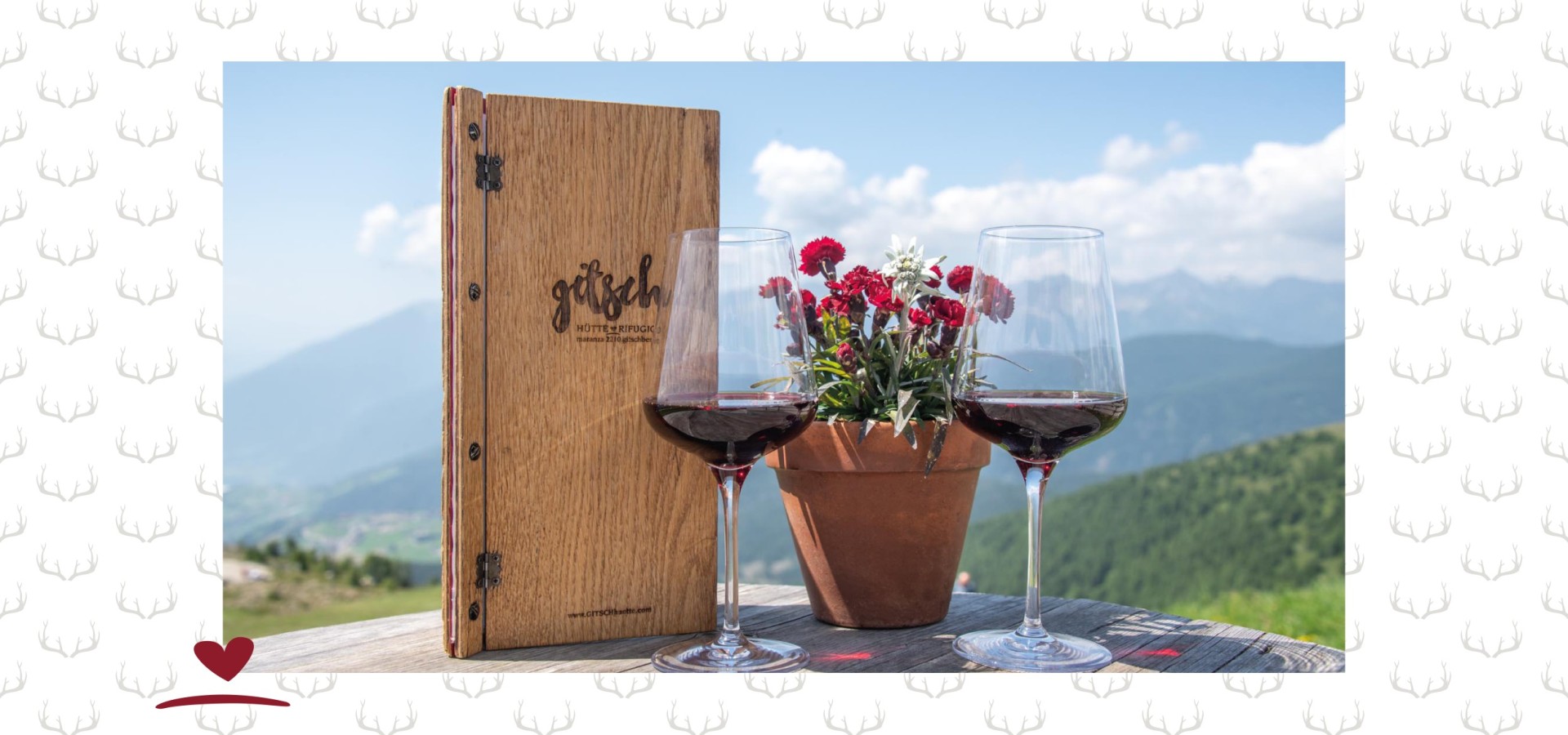 Menükarte aus Holz neben einem Topf mit Blumen und zwei Gläser Rotwein auf einem Holztisch. In Hintergrund Berge und blauer Himmel.