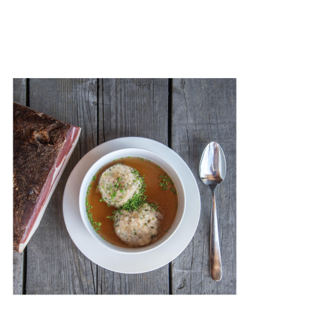 Ein Suppenteller mit zwei Knödel auf einem Holztisch. Daneben ein Löffel und ein Stück Speck.