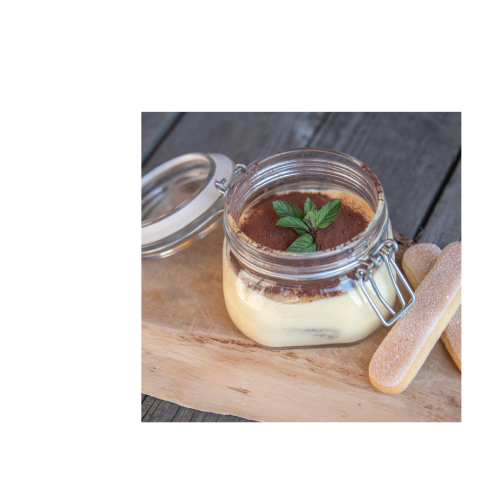 Ein Dessertglas gefüllt mit Tiramisu, Kakaopulver und Melisse auf einer Holzplatte. Daneben zwei Tiramisukekse.