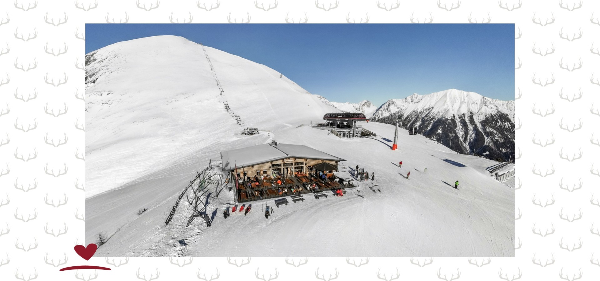 Bergrestaurant mit Terrasse und Sonnenschirmen auf schneebedecktem Berg, im Hintergrund schneebedeckte Gipfel und blauer Himmel.