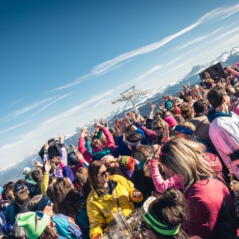 A lively crowd in colourful ski outfits at an outdoor party under blue skies with mountains in the background.