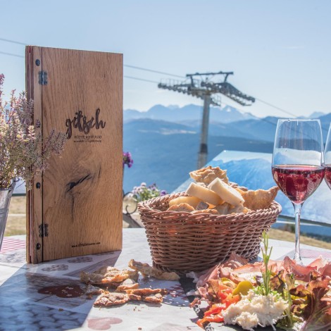 Outdoor table with a wooden menu, flowers, bread basket, sausage and cheese platter, and two glasses of red wine, with mountain scenery in the background.