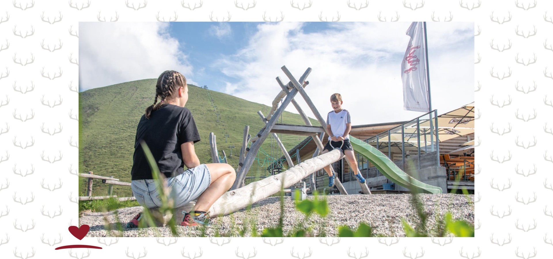 Kinder auf einer Holzwippe bei einem kleinen Spielplatz neben einer Hütte.