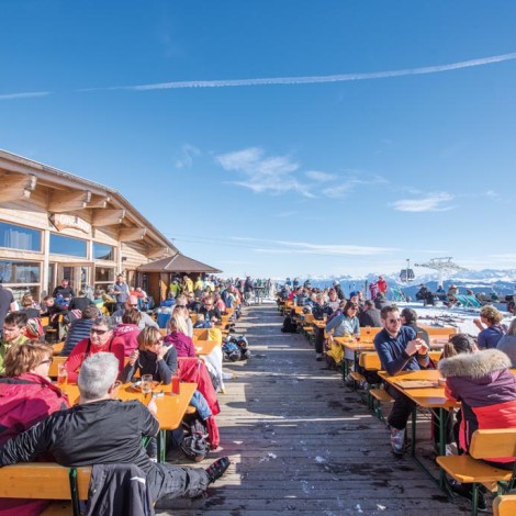 Terrace of a mountain restaurant with many customers seated at tables. In the background, snow-capped peaks and blue sky.