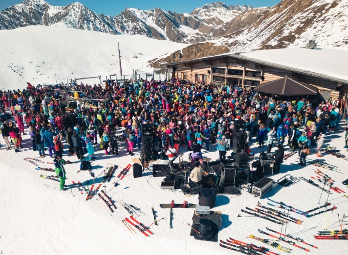 An open-air concert on a snow-covered mountain with a colourful crowd gathered in front of a mountain restaurant.