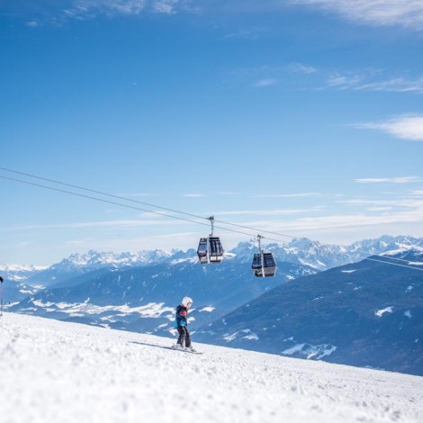 Two children skiing on the slopes, with two cable cars and a snow-covered mountain landscape under a blue sky in the background.