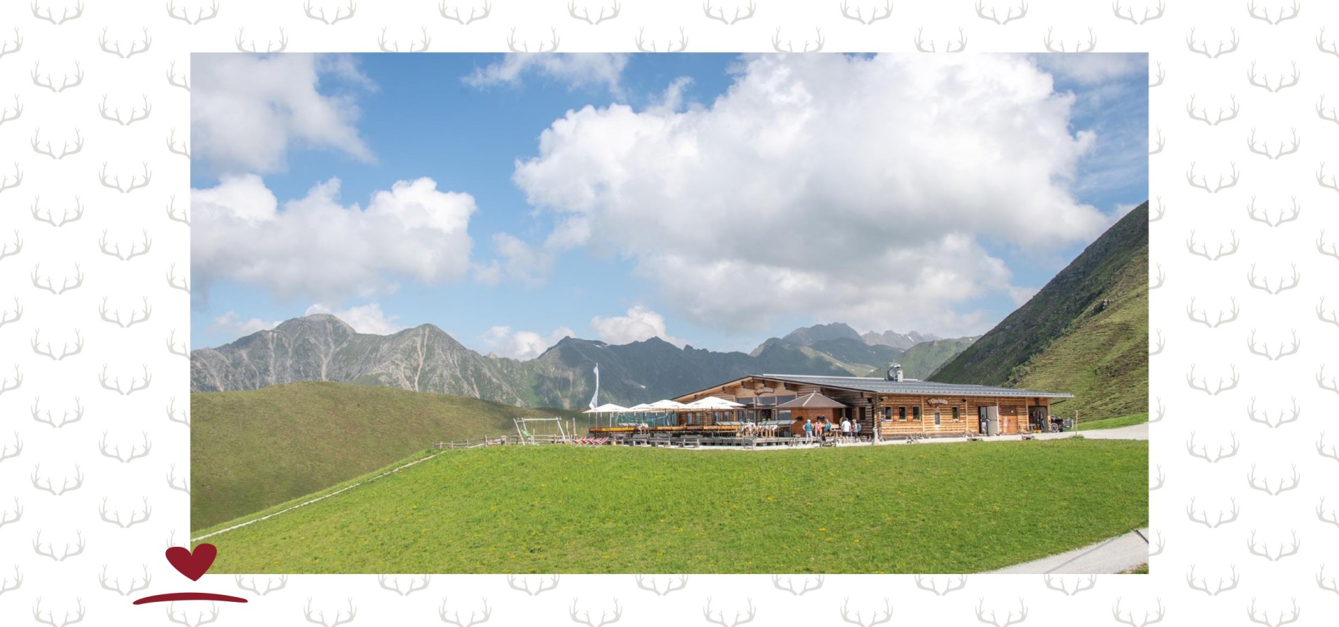 Bergrestaurant mit Terrasse umgeben von grünen Wiesen und einem Wanderweg. In Hintergrund ein Bergpanorama und ein leicht bewölkter Himmel.