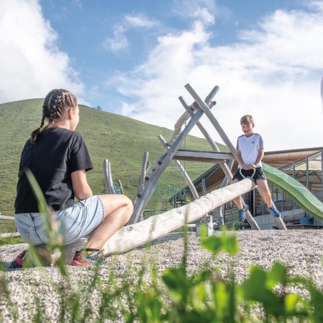 Two children play on a wooden seesaw in a playground with a mountain in the background.