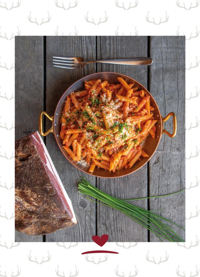 Eisenpfanne mit Nudeln und Tomatensauce auf einem Holztisch. Daneben eine Gabel, ein Stück Speck und Schnittlauch.