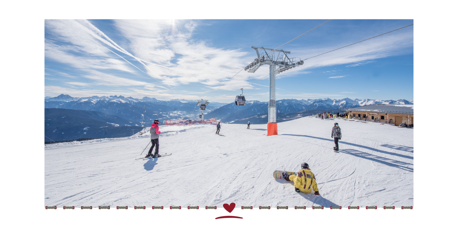 Skiers and snowboarders on the ski slope below the cable car. In the background, a mountain restaurant and snow-covered mountains under a blue sky.