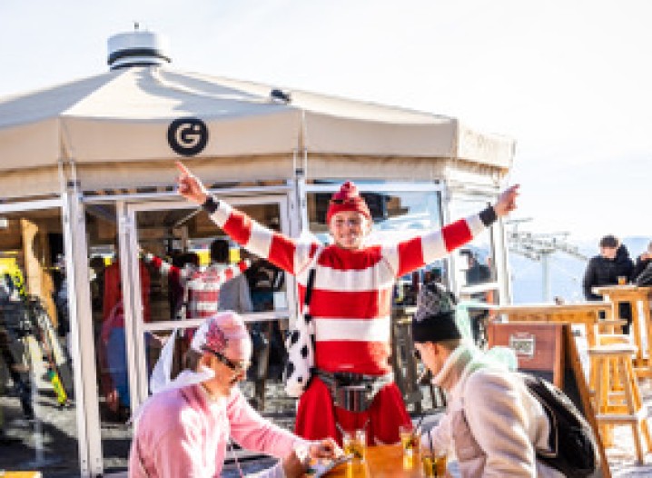 Person wearing a red and white striped jumper with arms raised in front of a curtain with a black “G” logo, other people seated at a table.