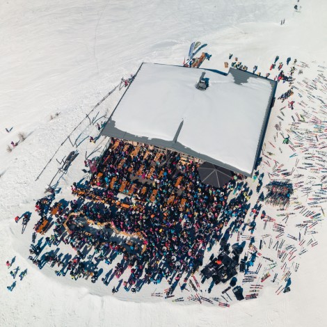 Aerial view of a crowd gathered next to a snowy landscape in front of a mountain restaurant. Skis are scattered on the right side of the hut.
