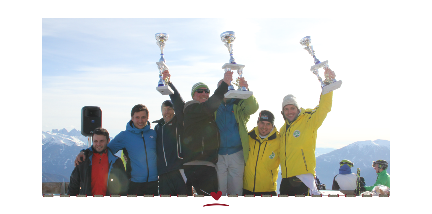A group of six people in winter clothing and smiling faces holding up three trophies. Snow-covered mountains and blue sky in the background.
