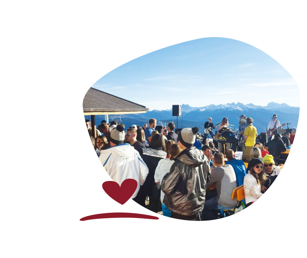 An event with lots of people and a band on a terrace in front of a mountain hut. In the background, snow-capped mountains under a blue sky.