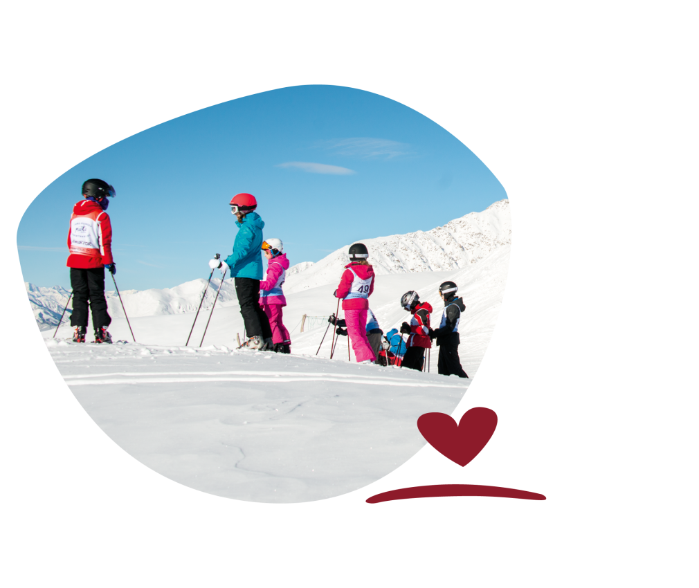Group of skiers in colourful clothing and helmets standing on a snow-covered slope in front of snow-covered mountains under a blue sky.