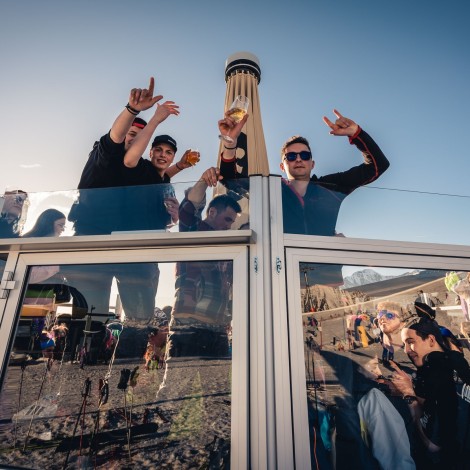 Three people are standing on a platform holding their drinks under a blue sky.